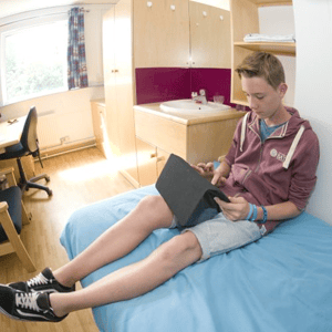 Un estudiante relajado en la cama de su habitación individual de la residencia de Bath College utilizando una tablet durante el curso de InterWay.
