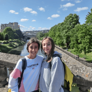 Dos estudiantes del curso de verano de InterWay en Bath posando juntas en un puente con un parque arbolado y el río de fondo en un día despejado.