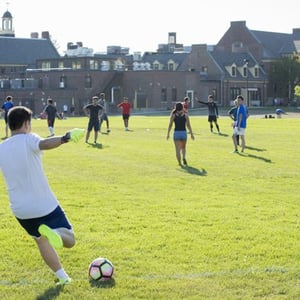 Un alumno del curso de inglés de InterWay en Boston chuta un balón de fútbol en un gran campo de césped exterior durante una actividad deportiva.