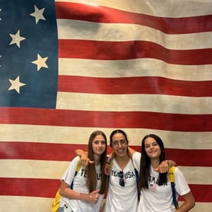 Tres estudiantes del viaje de idiomas de InterWay a Boston posando juntas delante de una enorme bandera de Estados Unidos.