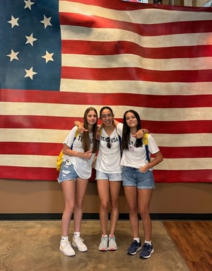 Tres jóvenes estudiantes del curso de verano de InterWay en EE. UU. posando abrazadas y sonrientes frente a una gran bandera americana de estilo rústico en interiores.