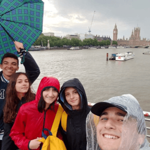Estudiantes del programa de idiomas de InterWay sonriendo con chubasqueros y paraguas junto al río Támesis, con el Big Ben y el Parlamento de Londres de fondo.
