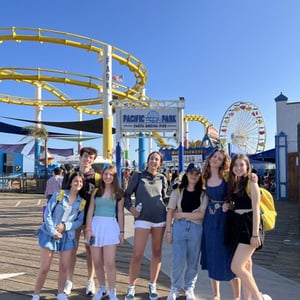 Cinco jóvenes alumnas del viaje de idiomas de InterWay posan sonrientes en un parque de atracciones de California con una montaña rusa amarilla de fondo.