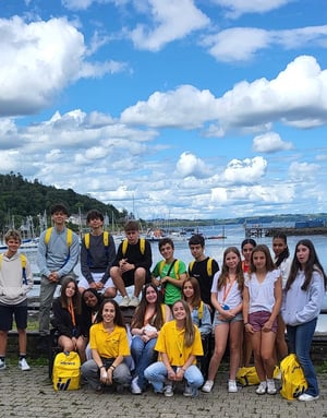 Gran grupo de estudiantes del curso de inglés en Cork College de InterWay posando juntos con mochilas amarillas junto a un puerto marítimo con barcos en Irlanda.