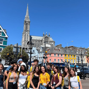 Alumnos de InterWay posando juntos durante una excursión cultural en el colorido pueblo costero de Cobh, Cork, con la catedral de San Colmán de fondo bajo un cielo despejado.