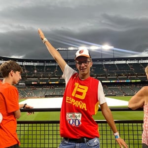 Un estudiante del programa de InterWay con camiseta roja anima con el brazo en alto en las gradas de un estadio deportivo en la Costa Este.