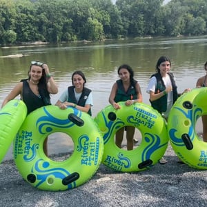 Cuatro estudiantes de InterWay posan sonrientes sosteniendo grandes flotadores verdes para hacer tubing en un río rodeado de árboles.