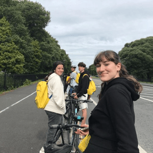 Una estudiante en primer plano sonríe durante una actividad de paseo en bicicleta con sus compañeros del curso Discover Dublin de InterWay, equipados con mochilas amarillas corporativas.