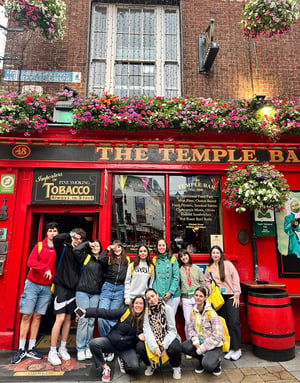 Grupo de estudiantes del curso de inglés en Dublín de InterWay posando sonrientes frente a la icónica fachada roja decorada con flores de The Temple Bar durante una excursión urbana.