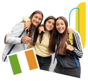 Tres estudiantes sonrientes de InterWay posando juntas con el pulgar hacia arriba durante un viaje de idiomas, con un gráfico de la bandera de Irlanda y un fondo de edificio tradicional de ladrillo.