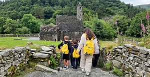 Alumnos de excursión con mochilas amarillas de InterWay subiendo unas escaleras de piedra hacia una antigua iglesia medieval con torre circular en Glendalough.
