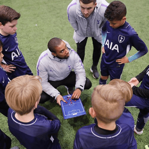Un entrenador con la equipación oficial del Tottenham Hotspur se reúne en círculo sobre el césped con los alumnos de InterWay para dar instrucciones tácticas con una pizarra.