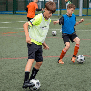 Estudiantes del campus de fútbol de InterWay en Londres practican el regate y el control de balón con botas de fútbol en un campo de césped artificial.