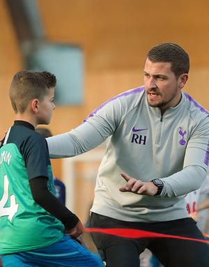 Un técnico de la academia del Tottenham con sudadera oficial da indicaciones motivacionales e individuales a un joven jugador del campus de fútbol de InterWay.