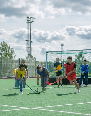 Alumnos del campamento de verano Hurst College Kids de InterWay jugando un partido de hockey sobre hierba con sticks en una pista deportiva exterior regulada.