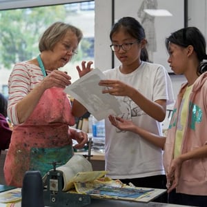 Una profesora nativa ayuda amablemente a dos alumnas del campamento Hurst College Kids de InterWay con un proyecto en papel dentro del aula.