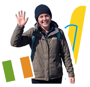 Una estudiante con gorro saludando en la naturaleza para el programa de InterWay en Irlanda, integrado con la bandera irlandesa y gráficos amarillos.