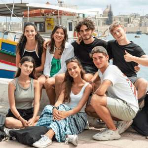 Grupo de alumnos de InterWay posando juntos y sonrientes en el paseo marítimo de Malta, con barcos tradicionales y la silueta de la ciudad histórica de fondo.