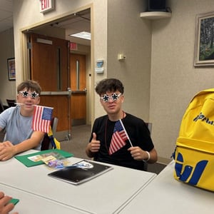 Dos alumnos del programa de idiomas de InterWay sentados en una mesa de clase con apuntes y una mochila escolar amarilla corporativa a su lado.