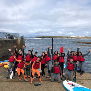Alumnos del programa de verano de InterWay equipados con neoprenos y chalecos salvavidas posan saludando junto a tablas de paddle surf en un embarcadero en Irlanda.