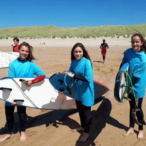 Tres jóvenes estudiantes con neoprenos de lycra azul sonríen en la arena sosteniendo tablas de surf durante una jornada de playa en el campamento de InterWay en Irlanda.