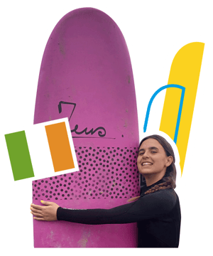 Una estudiante sonriendo mientras abraza una tabla de surf rosa durante el campamento multiactividad en Irlanda de InterWay, junto a la bandera irlandesa