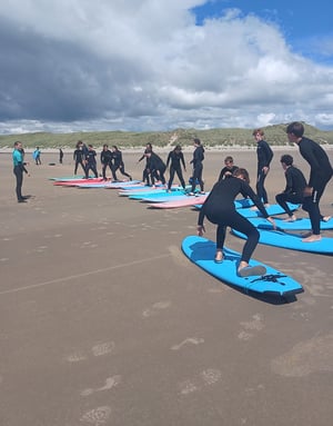 Grupo de estudiantes con trajes de neopreno negro practicando la puesta en pie sobre tablas de surf en la arena durante una clase del campamento de InterWay en Irlanda.