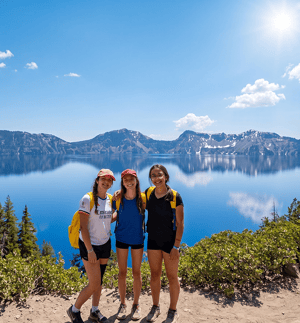 Tres alumnas del curso de inglés en Oregón de InterWay sonríen durante una excursión a Crater Lake, con las montañas nevadas y el agua azul cristalina reflejando el cielo de fondo.