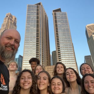 Un monitor y varias alumnas del programa de inglés de InterWay se toman un selfi sonriendo con dos altos edificios residenciales de fondo bajo un cielo azul claro.