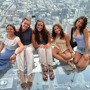 Cinco estudiantes de InterWay sentadas y sonriendo sobre el suelo de cristal transparente del mirador del Arco de St. Louis con vistas urbanas elevadas bajo sus pies.