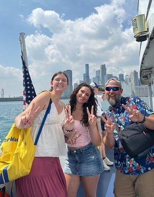 Dos alumnas con una mochila amarilla y un monitor de InterWay posan sonrientes a bordo de un barco con banderas americanas y el horizonte urbano al fondo.