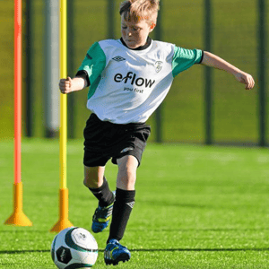 Un niño con equipación deportiva blanca y verde regatea con el balón entre picas de entrenamiento naranjas y amarillas en el Soccer Camp de InterWay en Irlanda.
