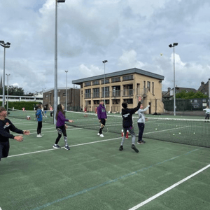 Grupo de estudiantes participando en una clase colectiva de tenis en pistas de césped sintético al aire libre con el edificio del club al fondo, una de las actividades del campus de InterWay.