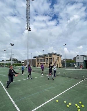 Un grupo de jóvenes estudiantes del programa deportivo de InterWay entrena con raquetas y pelotas en una pista de tenis exterior en Irlanda bajo un cielo nublado.