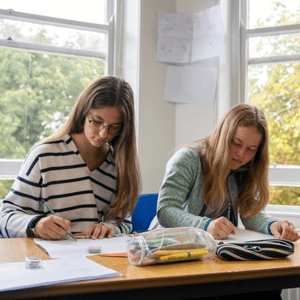Dos estudiantes del curso de inglés en Torquay de InterWay concentradas escribiendo en sus cuadernos durante una clase junto a la ventana.