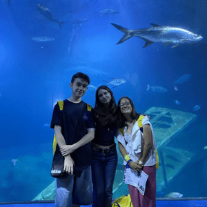 Tres jóvenes estudiantes del viaje de InterWay en Torquay posando sonrientes frente al gran tanque de agua azul de un acuario con peces de fondo.
