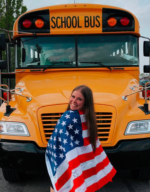 Alumna de Interway envuelta en la bandera estadounidense frente al típico autobús escolar amarillo